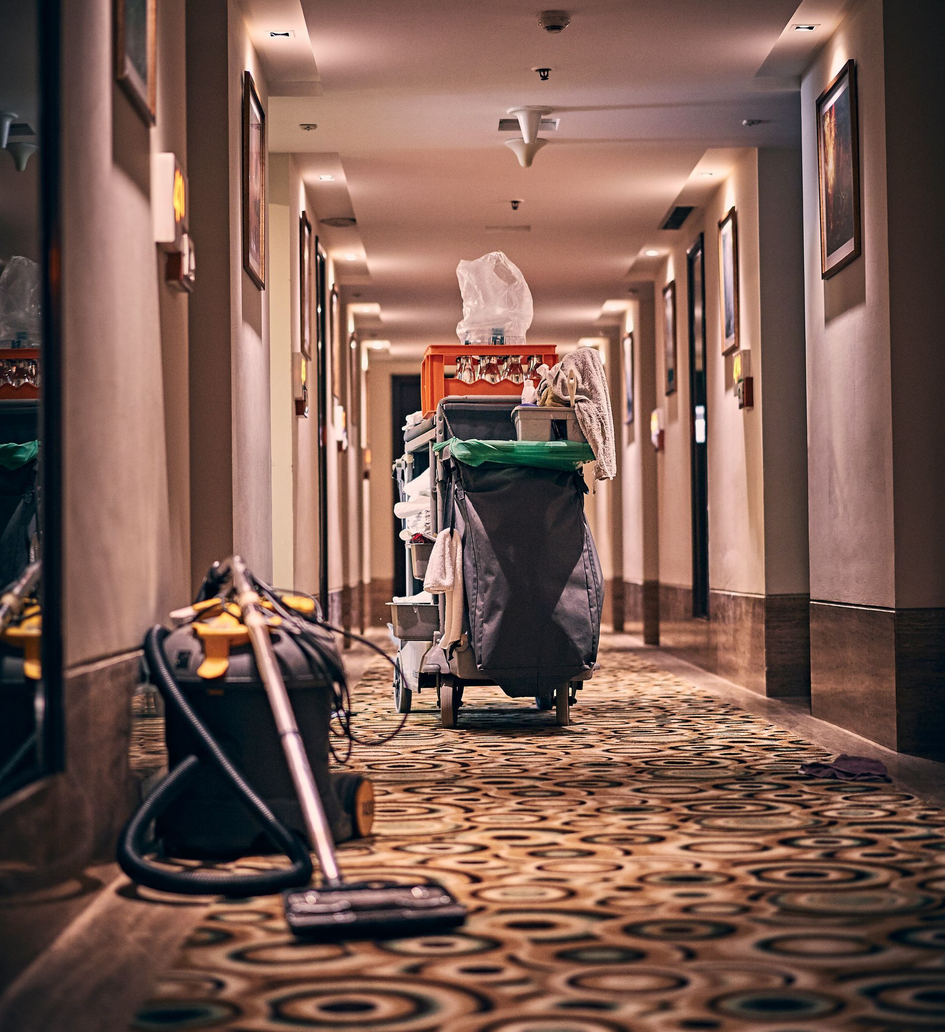A housekeeping cart and vacuum cleaner in a long hotel corridor with patterned carpet